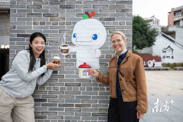 來(lái)自英國(guó)的游客（右）手持十五運(yùn)特色杯子與喝咖啡的“喜洋洋”墻繪合影。